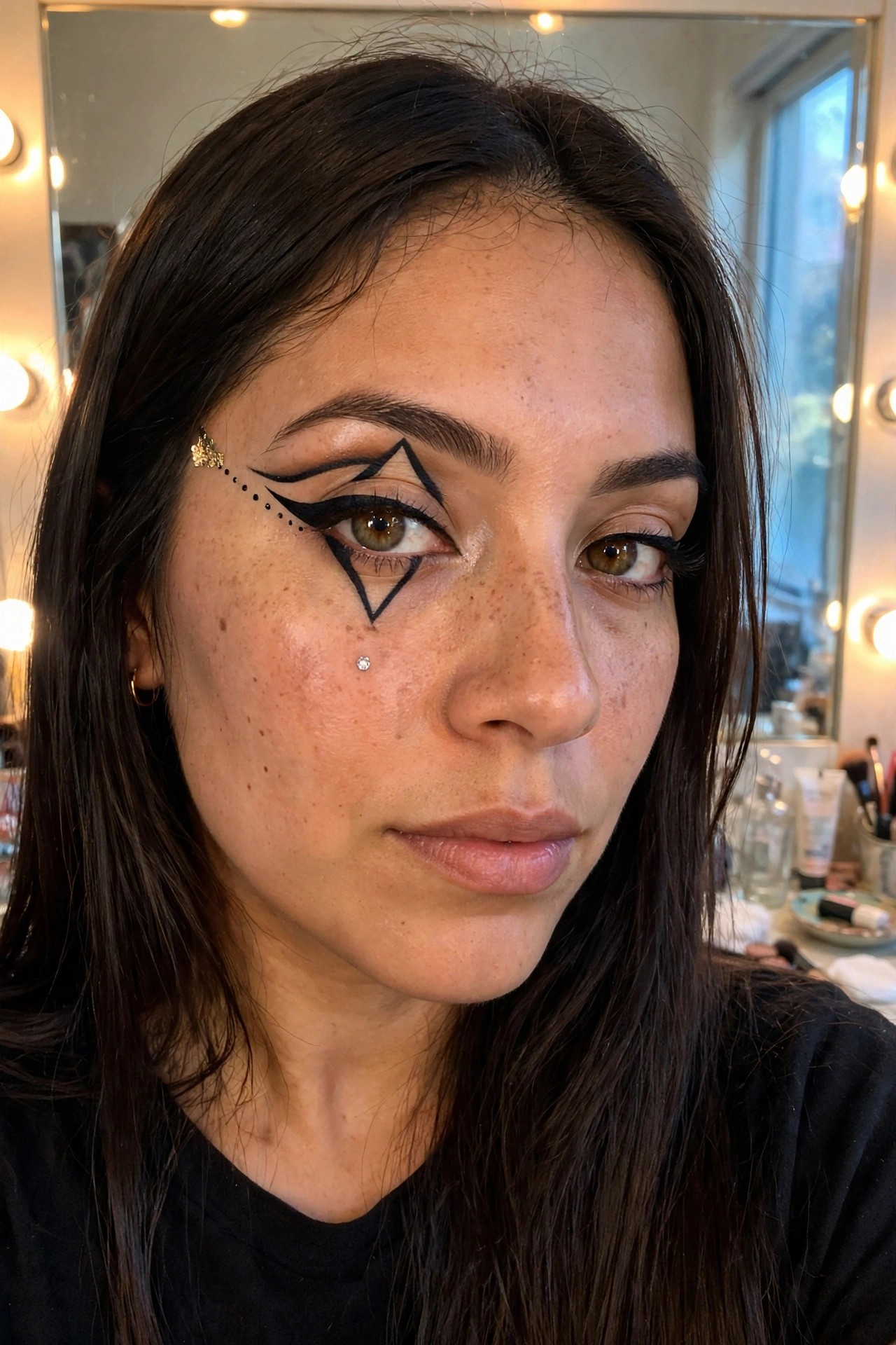 Close up of a woman with long dark hair wearing bold black graphic eyeliner in triangular shapes around one eye with dotted lines extending outward and small rhinestone and gold star accents.