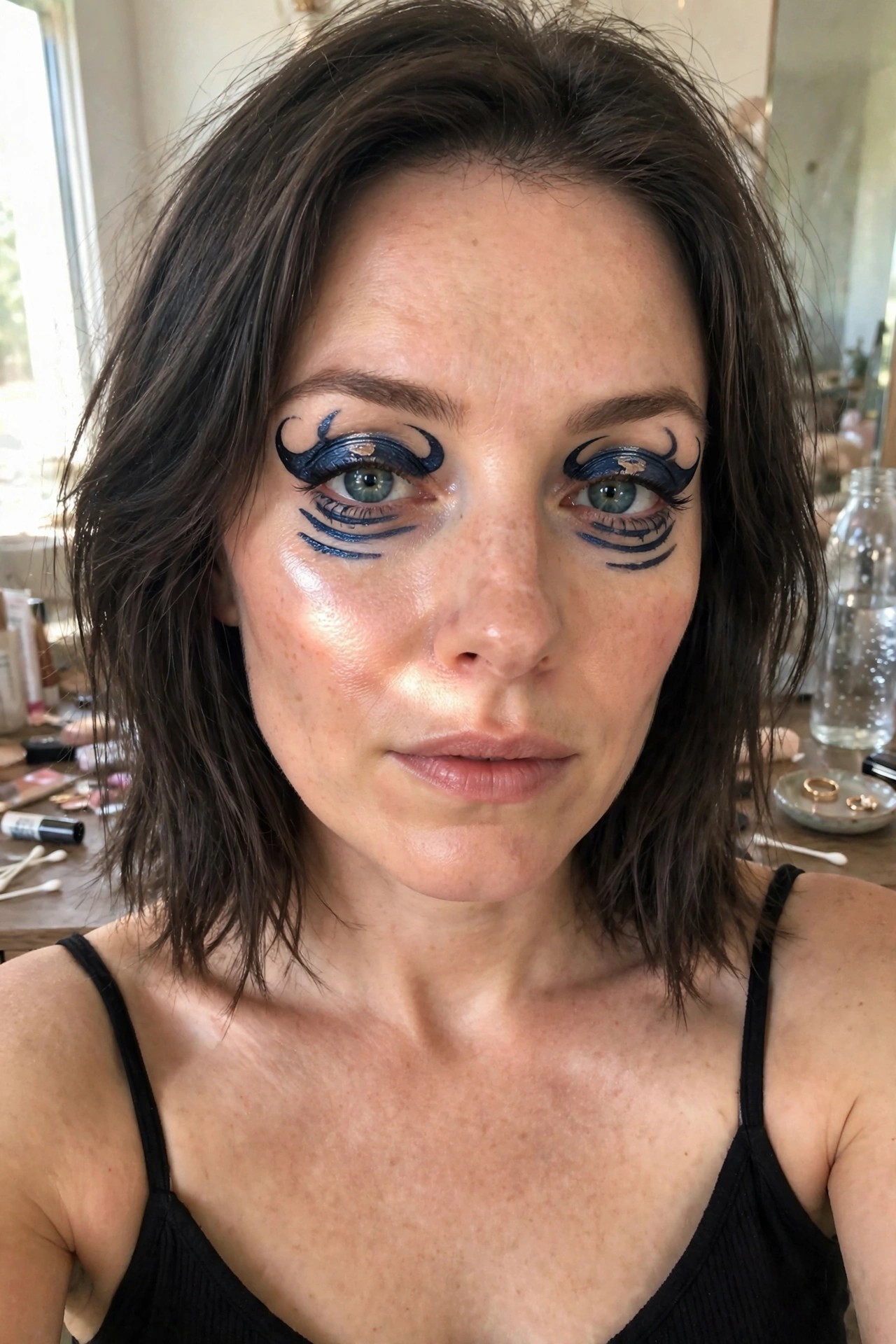 Close-up of a woman with short brown hair showing bold blue and black graphic eyeliner featuring swirls above the eyes and multiple extending lines below each eye.