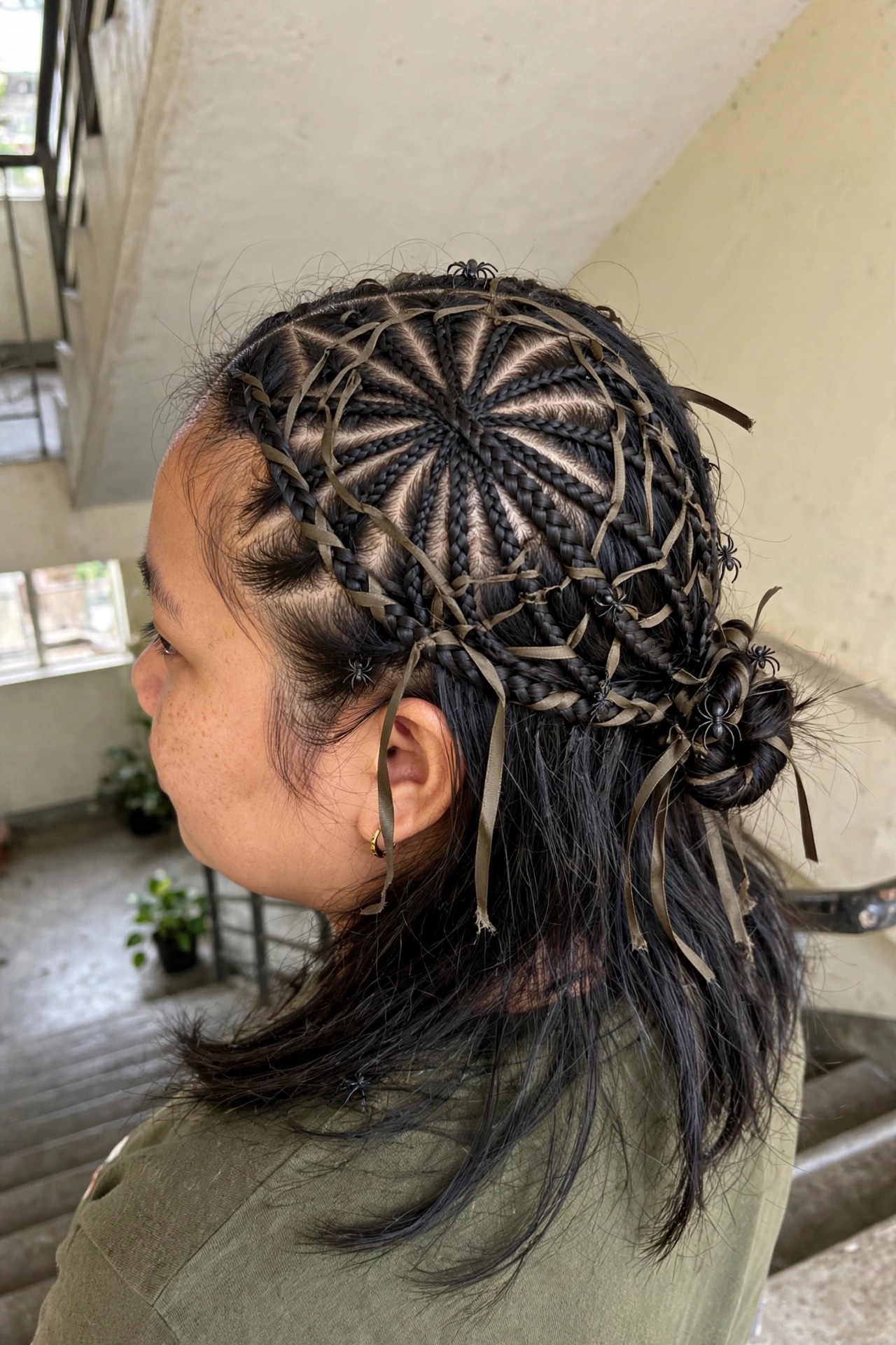 Close side view of a woman wearing an intricate radial braided updo with olive ribbons woven through the braids to form a spiderweb pattern and small black spider accents placed throughout the style.