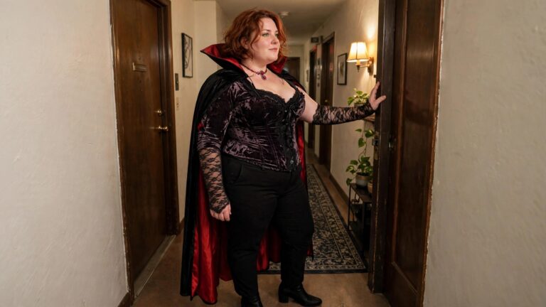 18 Trendy Plus Size Halloween Looks for Confidence and Style Plus size woman wearing a black velvet vampire corset with red lined cape, black pants, black boots, and lace gloves while standing in a hallway.