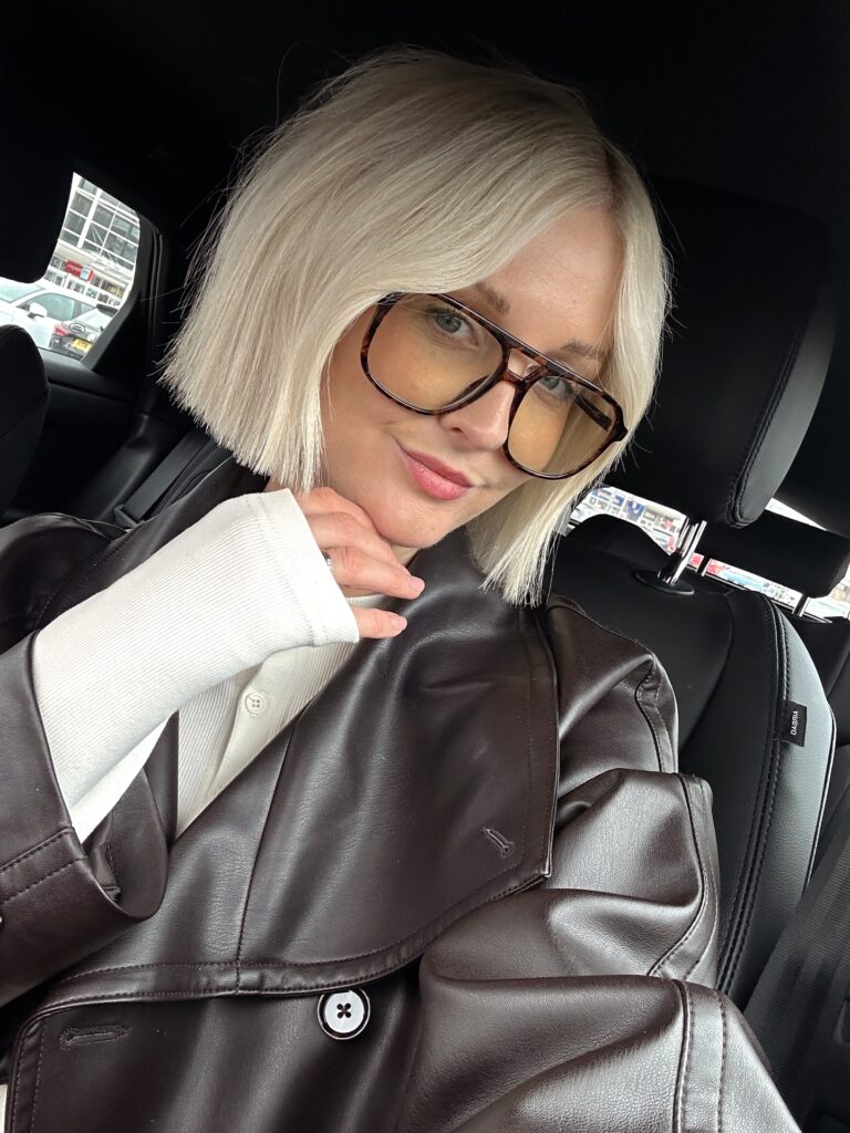 A woman with a chin-length, blunt-cut platinum blonde bob poses in a car wearing glasses.
