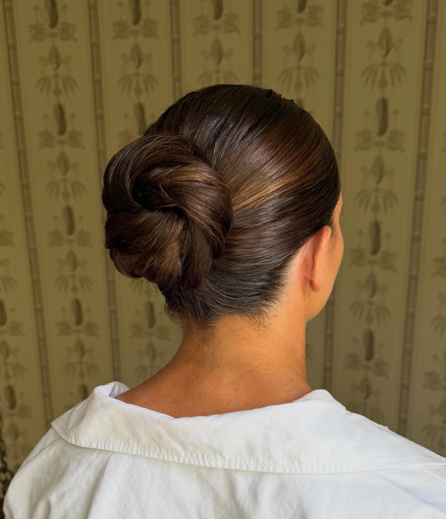 Back view of a woman with glossy chocolate brown hair and caramel highlights in a sleek slicked-back low bun with a twisted rosette shape.