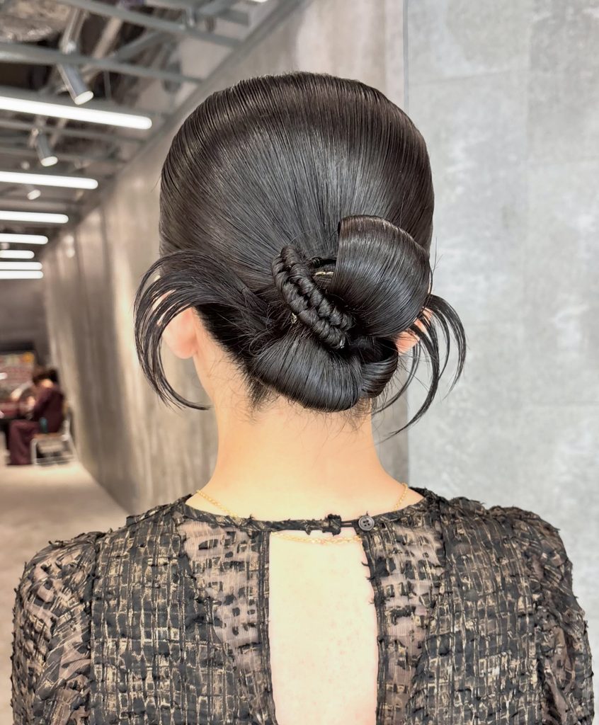 Back view of a woman with jet black hair in a glossy slicked-back low sculptural bun with a braided detail and thin strands framing the ears.