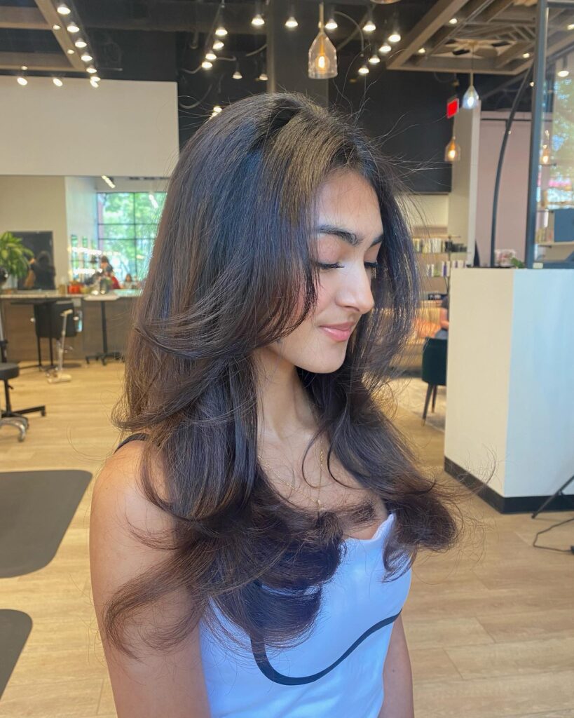 A woman with long dark brunette hair featuring voluminous roots and soft curled under layers.
