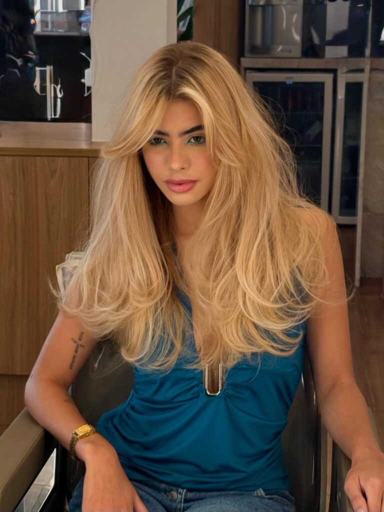 A woman with long golden blonde hair featuring feathered layers, a center part, and soft cascading waves.
