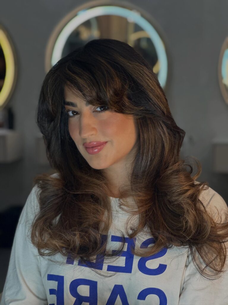 A woman with long brunette hair featuring caramel highlights, voluminous curled ends, and wispy bangs.