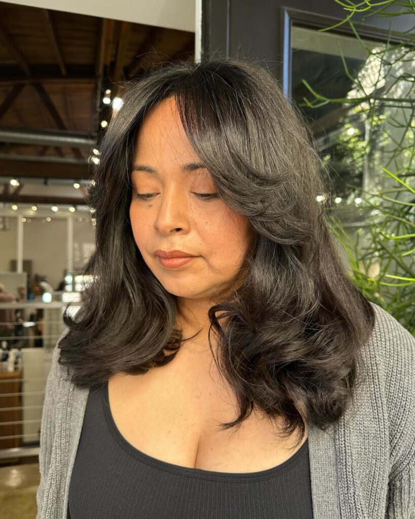 A woman with medium length dark brunette hair featuring curled-under layers, natural gray strands, and long side swept bangs.