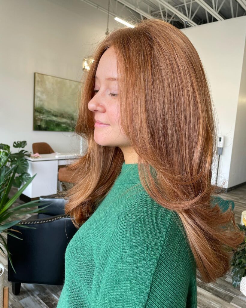 A woman with long copper red hair styled in soft face-framing layers with flipped-out ends and a subtle curtain part.