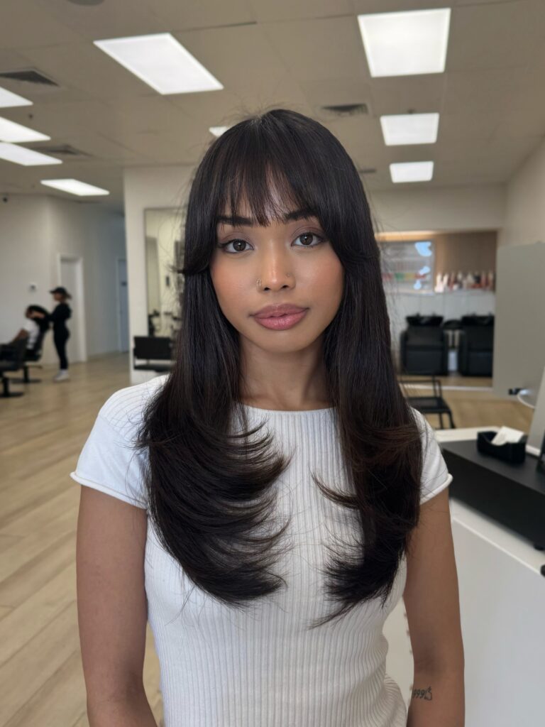 A woman with long black hair featuring full blunt bangs and face framing layers that flip outward.