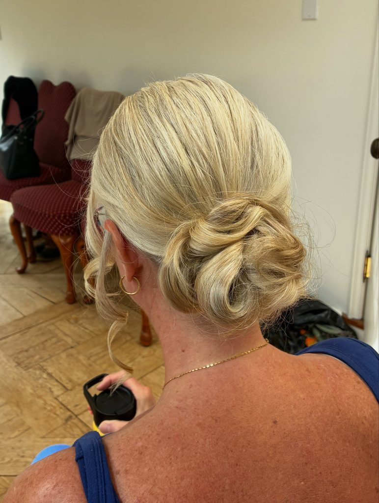 Back view of a woman with golden blonde hair styled in a soft twisted low bun with curled tendrils near the ear and a small gold hoop earring.