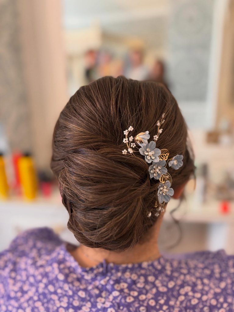 Back view of a woman with chestnut brown hair styled in a sleek low French twist with a blue floral hair comb with pearls and gold leaves.