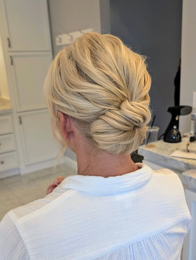 Back view of a woman with bright blonde highlighted hair styled in a soft twisted low bun with a swept-back crown and a few wispy strands near the ear.