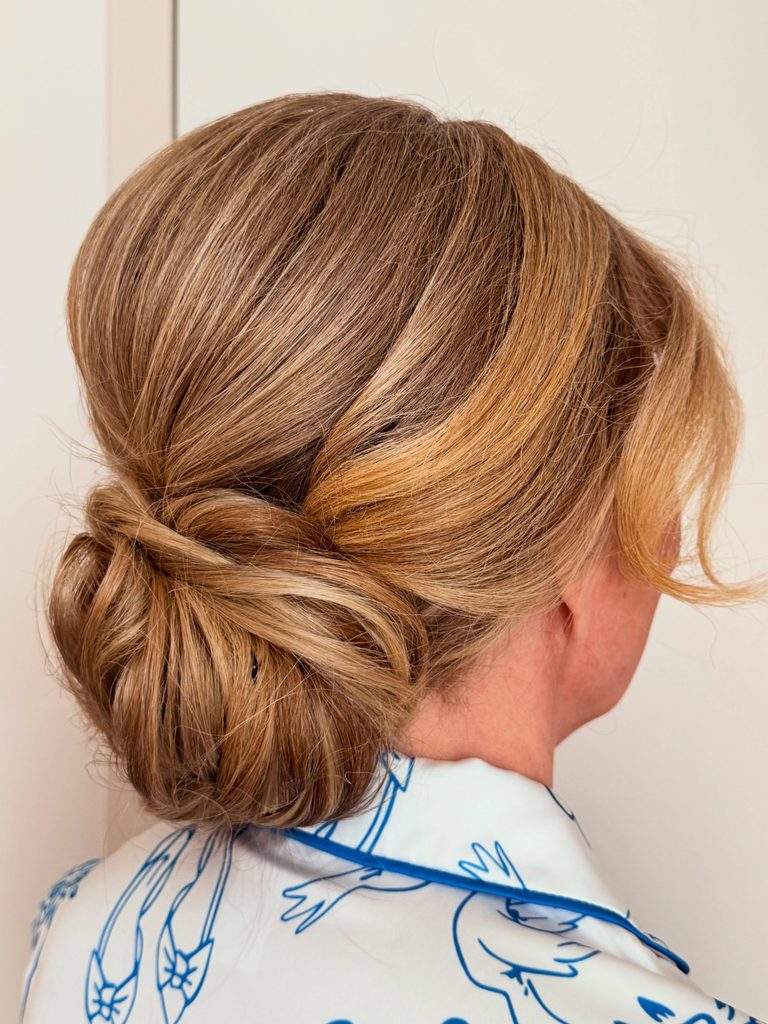 Back view of a woman with caramel blonde highlighted hair styled in a soft low chignon with a voluminous crown and a loose wispy strand at the side.