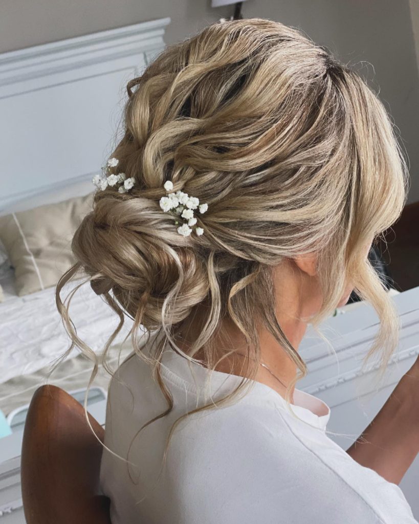 Back view of a woman with dark blonde highlighted hair styled in a romantic wavy low bun with loose tendrils and small white baby's breath flowers.