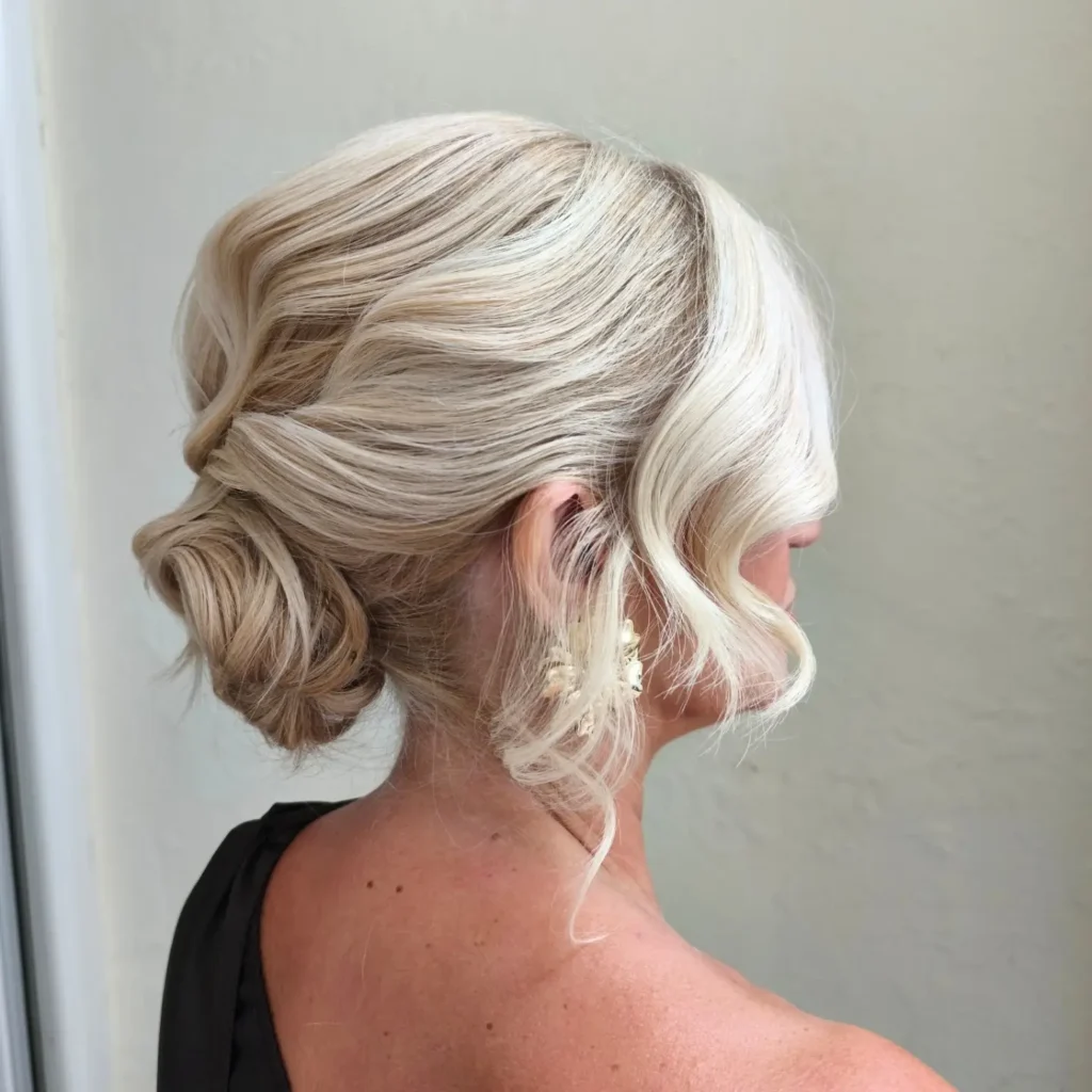 Side view of a woman with icy platinum blonde hair styled in a soft twisted low bun with a wavy face-framing piece and a floral drop earring.