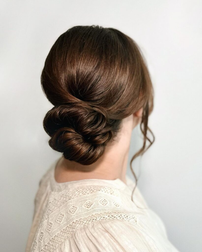 A woman with glossy dark brown hair styled into a smooth twisted low bun with a single wavy side tendril, seen from behind wearing a cream lace top.