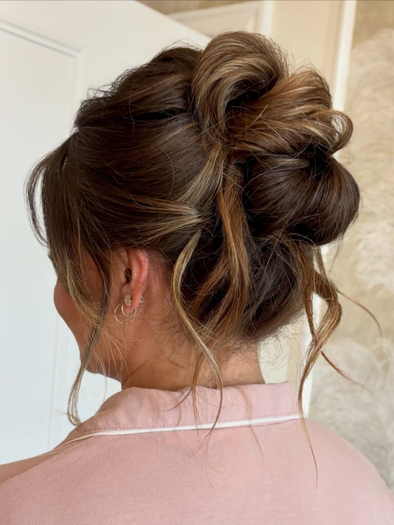  A woman with brunette and golden highlighted hair in a tousled high updo with loose wispy tendrils around her ears, shown from behind wearing a pink top.