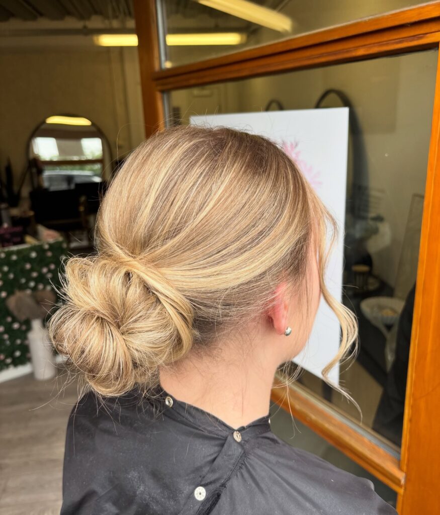 A woman with sandy blonde highlighted hair styled into a low wrapped bun with soft face framing curls, shown from a side profile in a salon setting.