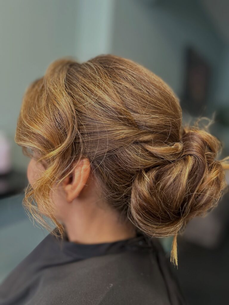 A woman with warm caramel brown hair with golden highlights styled into a relaxed low chignon with swept back side layers, shown in profile.