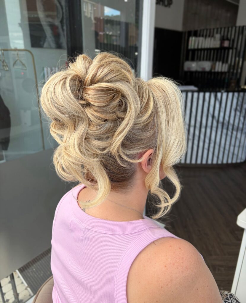 A woman with bright blonde highlighted hair in a high voluminous updo with curly loops and sweeping side bangs, shown from the side back in a pink top.