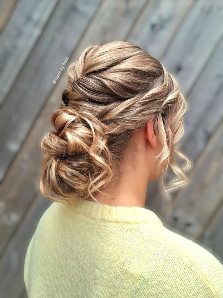 A woman with wavy blonde highlighted hair styled into an elegant, low, textured bun with loose tendrils, seen from the back, wearing a light yellow sweater against a rustic wooden fence background.