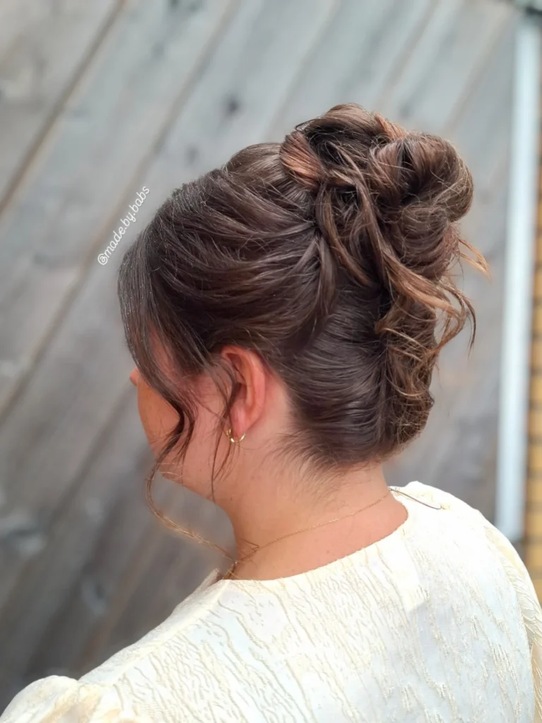 A woman with brown hair in a textured high updo with soft curls on top and face framing tendrils, shown from a side profile.