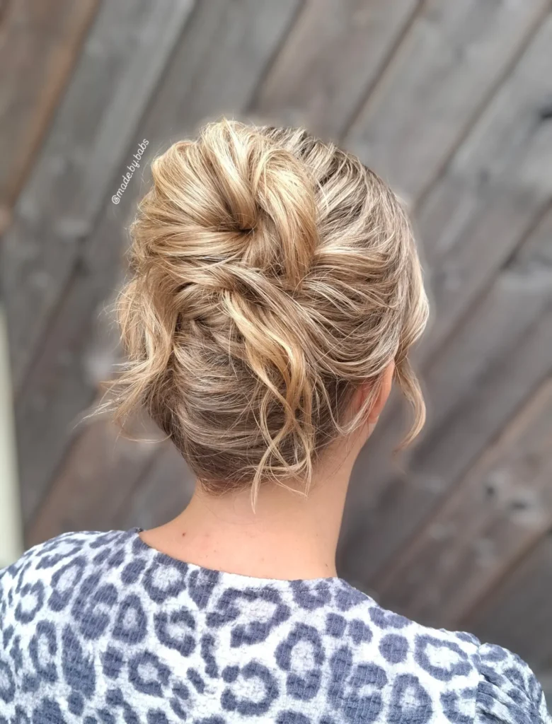 Back view of a woman with blonde highlighted hair styled in a twisted messy bun with loose wavy strands at the nape.