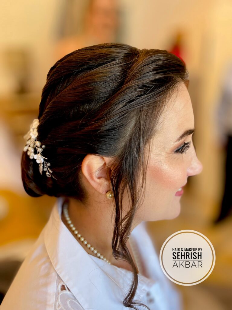 Side view of a woman with dark brown hair styled in a sleek low twisted chignon with a pearl and flower hair vine and one loose strand near her face.