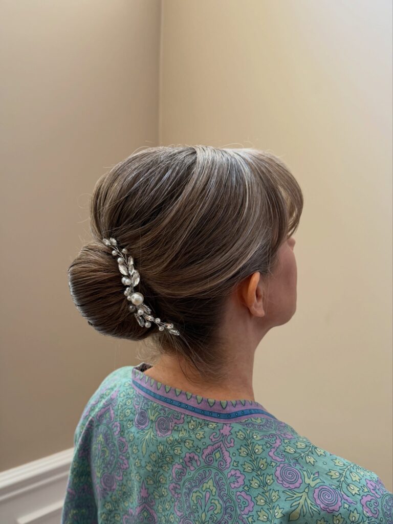 Back view of a woman with brunette and silver highlighted hair styled in a smooth low bun with a voluminous crown, side swept bangs, and a crystal and pearl hair vine.