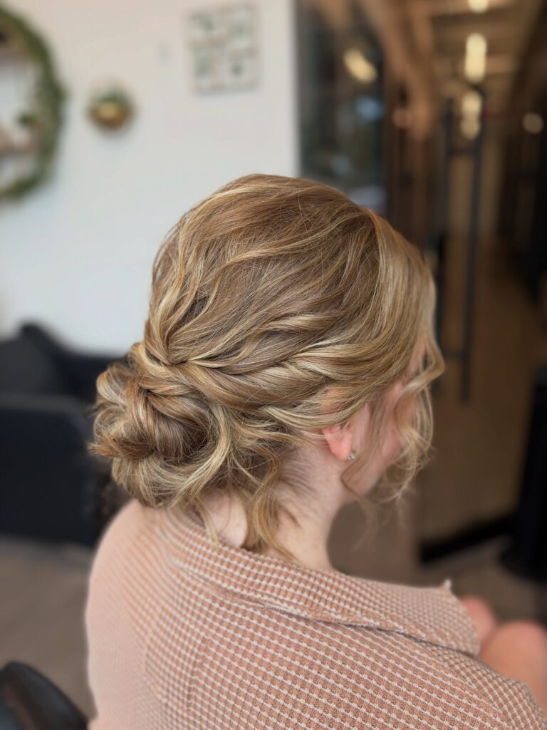Back view of a woman with honey blonde highlighted hair styled in a soft low bun with rope twists and loose waves near her ears.