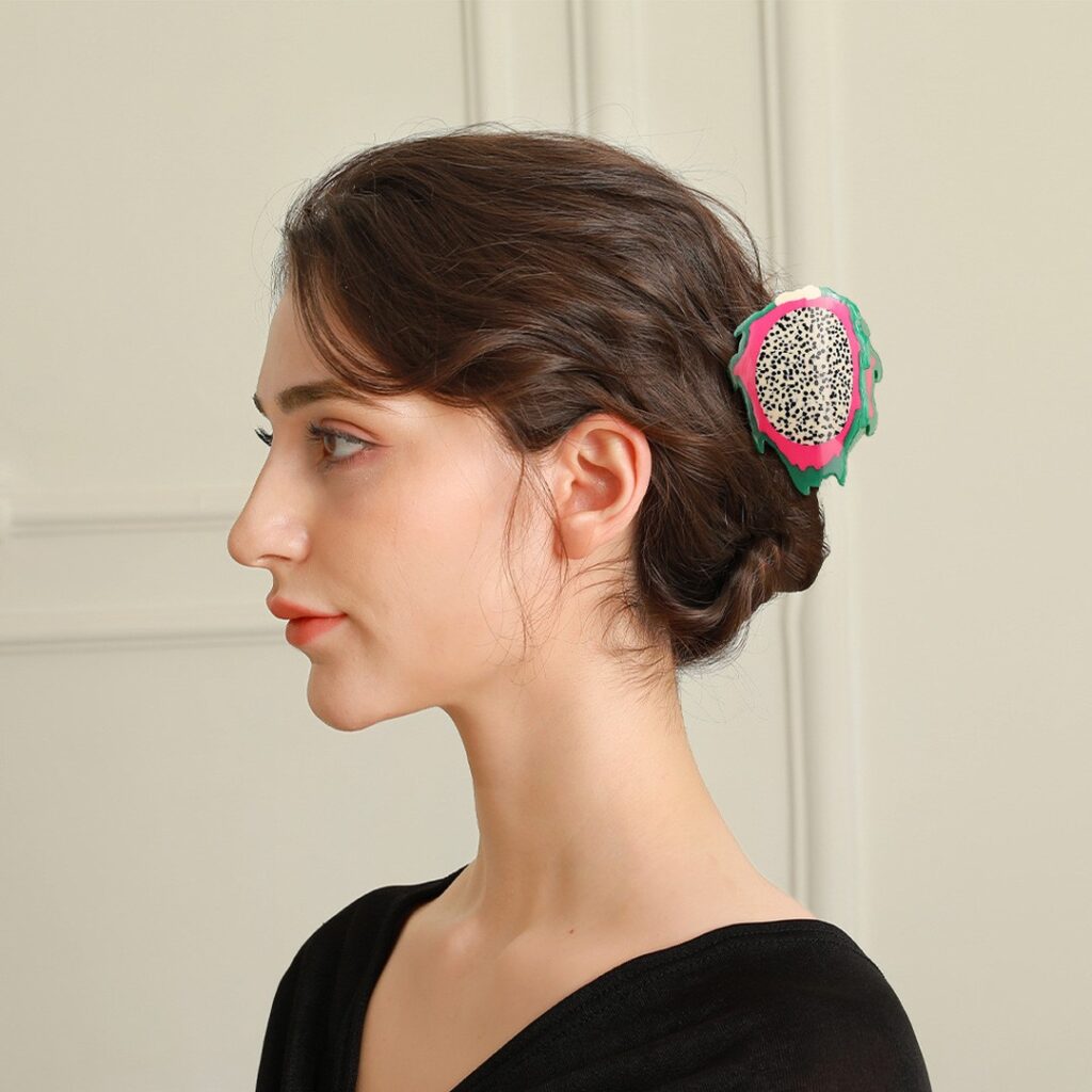 A woman with dark wavy hair wears a twisted low bun accented with a bold pink and green dragon fruit shaped hair clip.