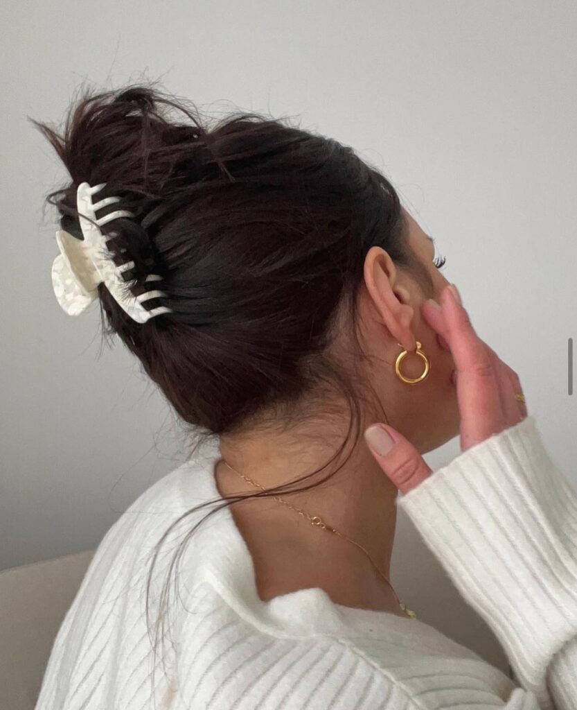 A woman with dark brunette hair wears a loose, textured top bun secured with a cream marbled claw clip and face framing wisps.