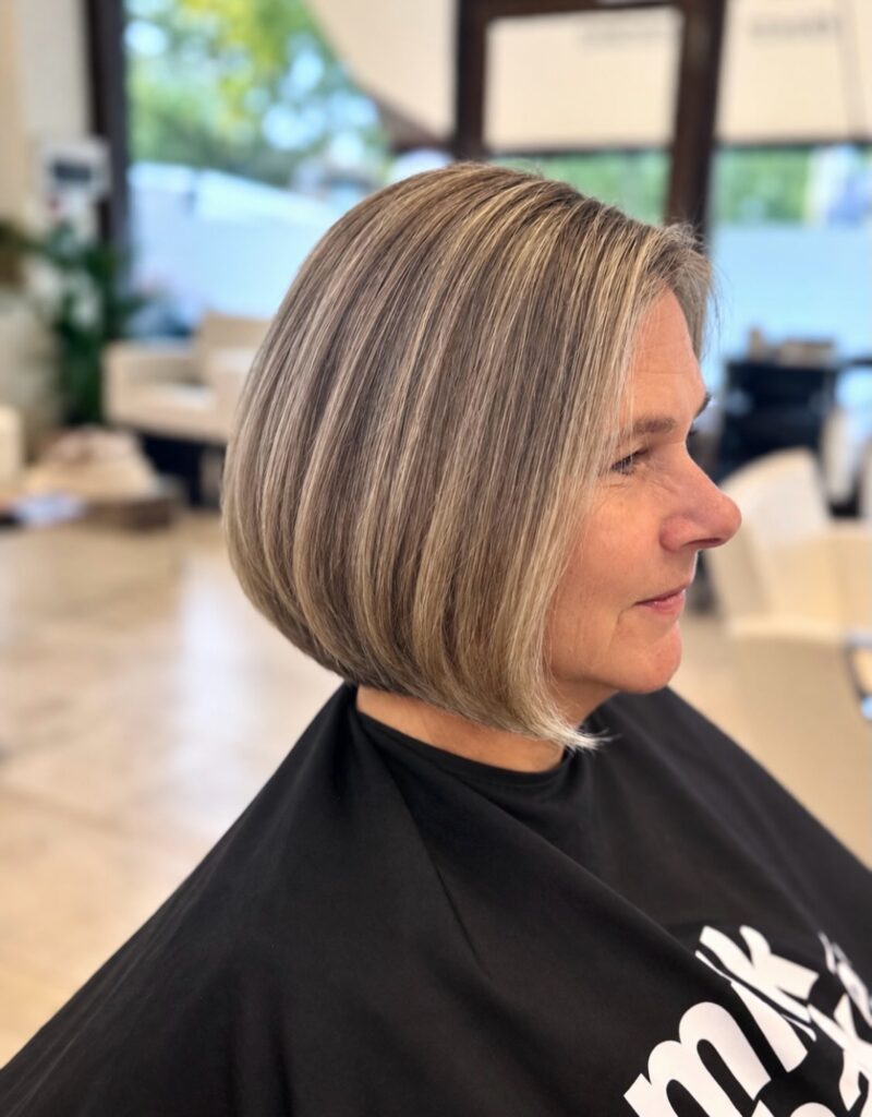 A woman with a rounded angled bob featuring soft brown and blonde highlights is shown in profile at a salon.