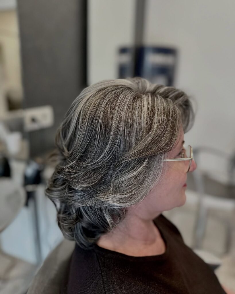 A woman with medium length blown out waves in a natural grey and dark blend is shown in profile wearing glasses.