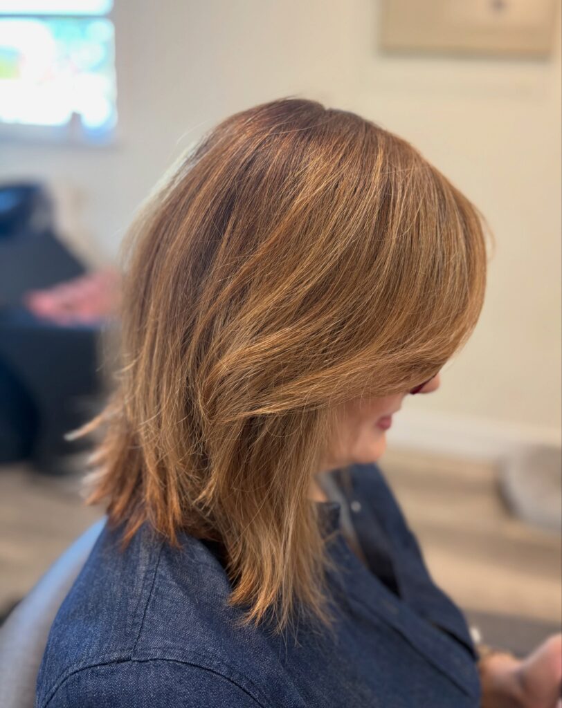A woman with shoulder length layered hair in warm caramel tones and a curtain fringe is shown in profile.