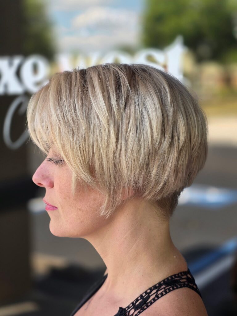 A woman with a short stacked wedge bob in dimensional blonde is shown in profile against a storefront window.
