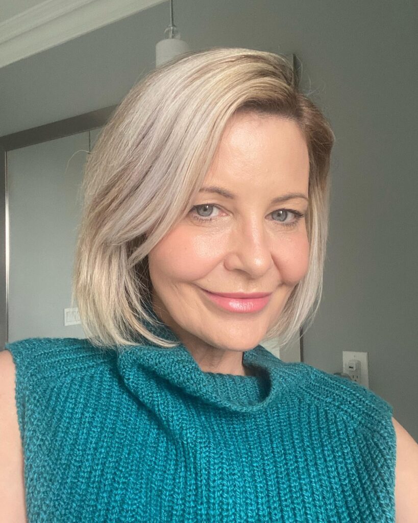 A woman with a sleek chin-length blonde balayage bob and a deep side part smiles while wearing a teal sweater.
