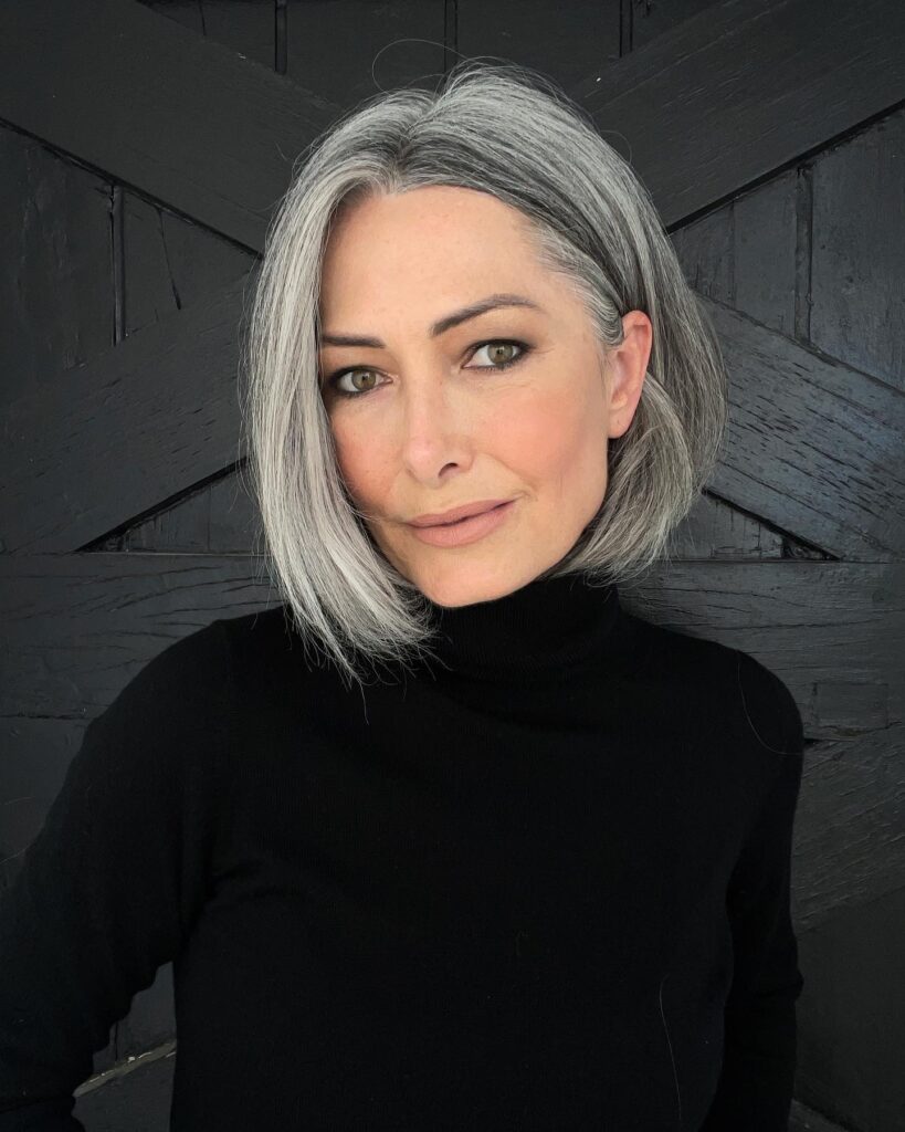 A woman with a straight silver bob and a center part wears a black turtleneck in front of a dark wooden wall.