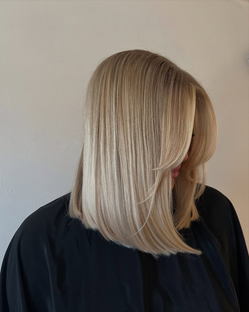 A woman with a sleek, straight buttery blonde lob featuring subtle highlights is shown from the side.