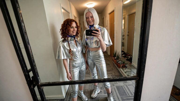 21+ Bold Matching Halloween Costume Ideas That Turn Heads Two women in matching shiny silver futuristic jumpsuits with glowing blue LED collars, short capes, and silver boots taking a mirror selfie.