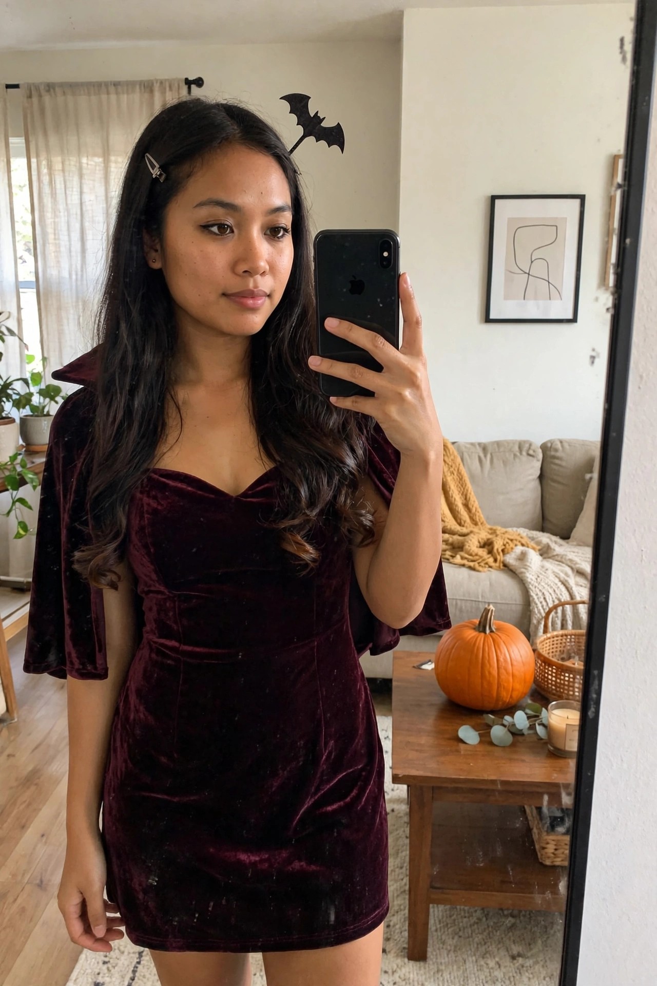 Woman wearing a short burgundy velvet dress with wide structured collar and cape sleeves, long dark hair with a black bat clip, holding a phone for a mirror selfie.