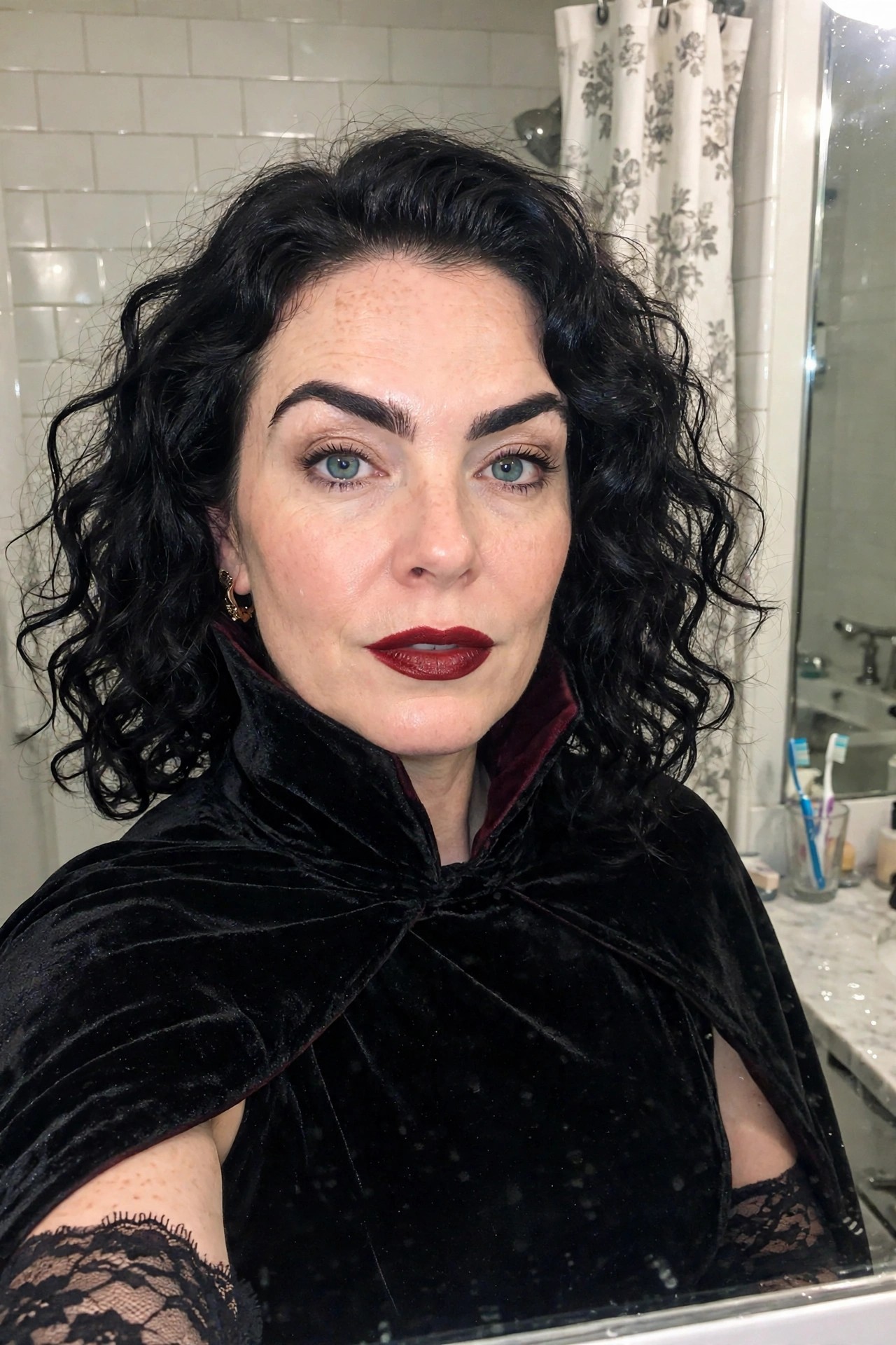Woman wearing a black velvet cape with red lining and a high collar paired with dark red lipstick and voluminous black curly hair.