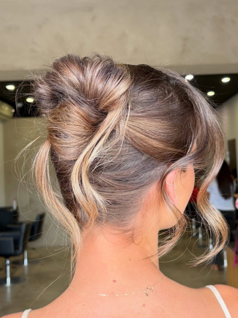 A woman with thick balayage hair wears a loose twisted low bun updo with face-framing pieces left out.