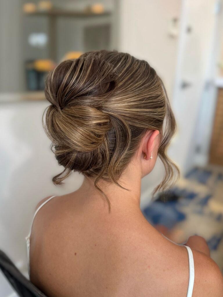 A woman viewed from behind has her thick highlighted hair styled in a sleek French twist with soft curled face framing strands and a few tendrils.