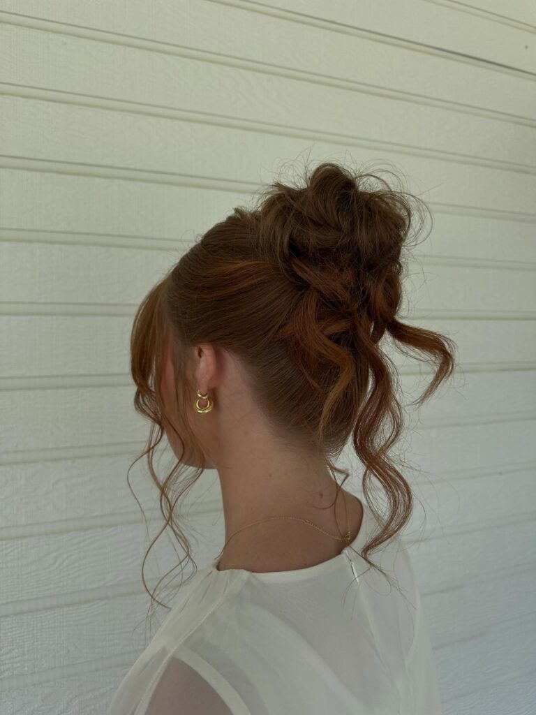 A woman with auburn hair wears a tousled high bun updo with loose curly tendrils framing her face.