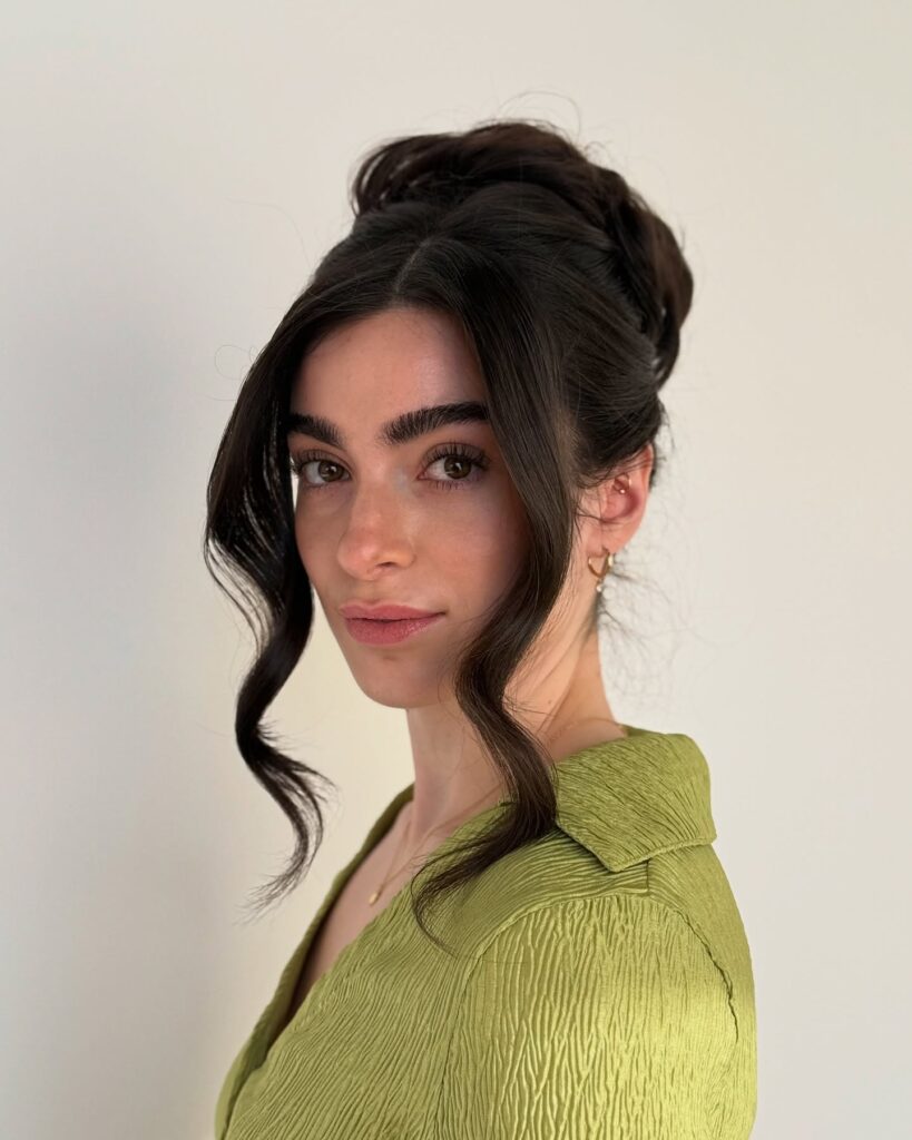 A woman with long dark chestnut hair wearing a textured high bun with two long curled tendrils framing her face.
