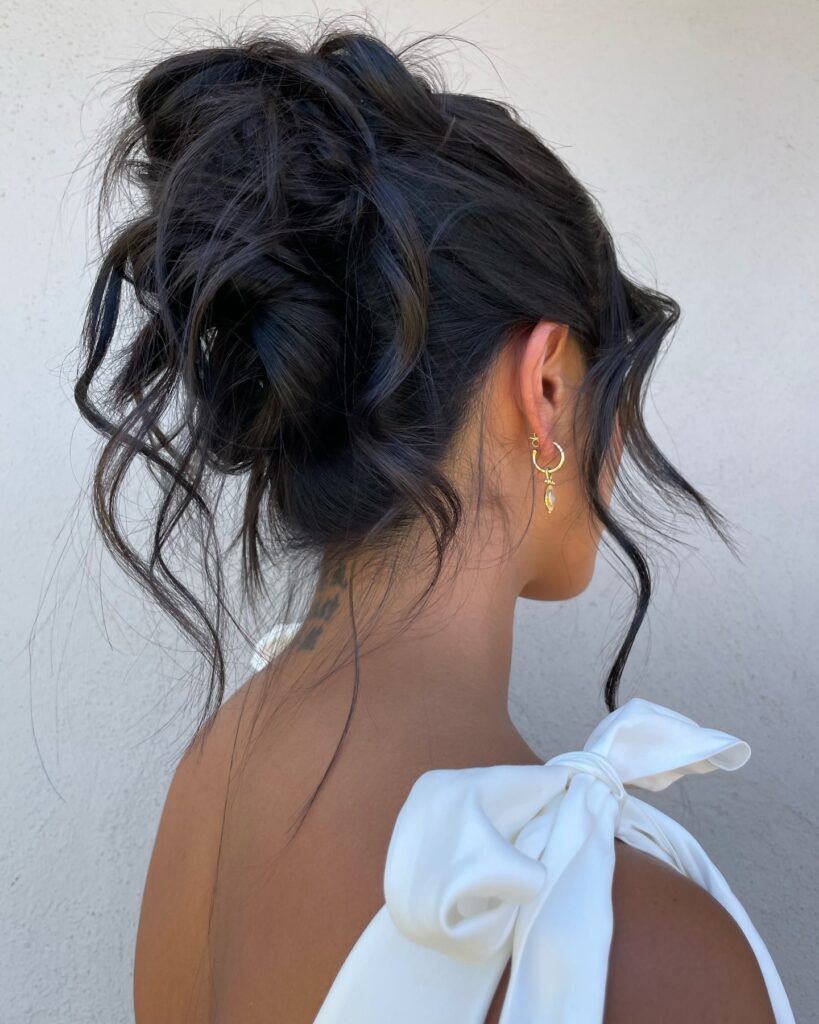 A woman viewed from behind with her thick, dark hair styled in a messy, textured updo with wavy strands framing her face and neck.