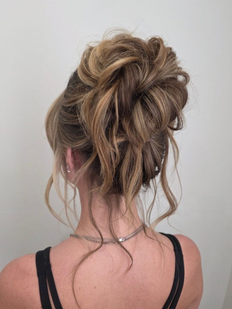 A woman with medium length highlighted hair styled in a high, messy bun with loose wavy face framing tendrils and strands on the nape.