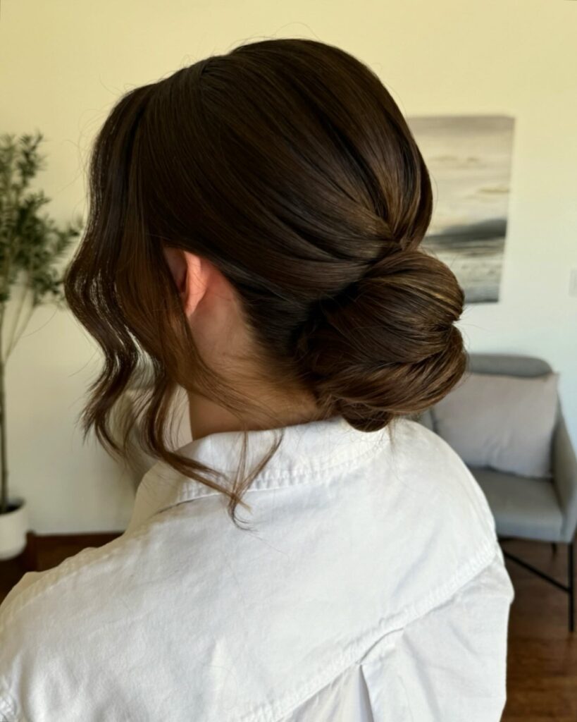 A woman with thick brunette hair styled in a sleek low twisted chignon with soft wavy tendrils framing her face.