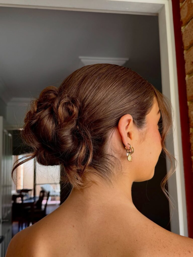 A woman with chestnut brown hair wears a low rose-shaped bun updo with soft face-framing strands.